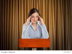 Panische Angst vor Präsentationen. Das Bild zeigt eine Frau vor einem Rednerpult mit 2 kleinen Mikrofonen. Sie stützt ihren Kopf mit beiden Händen und starrt ins Leere.