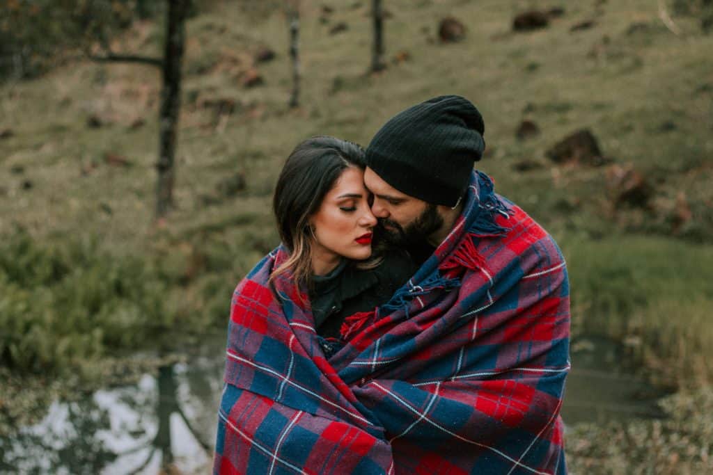 Beziehungscoach: auf dem Foto ist ein Paar zu sehen, das eng umschlungen unter einer Wolldecke an einem grünen Bergabhang steht.