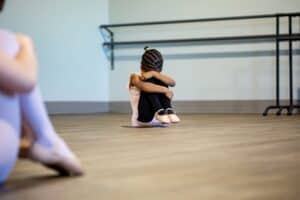 Bühnenangst: Auf dem Bild ist ein kleines Mädchen in Ballettkleidung zu sehen. Sie sitzt auf dem Boden, der Kopf ist auf den Kopf gesenkt und durch ihre Arme geschützt. Es scheint, sie hat Angst oder ist demotiviert.