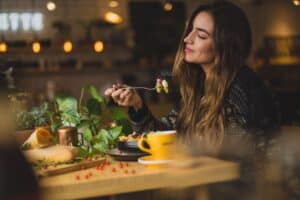 Abnehmen durch Psychologie. Auf dem Bild ist eine junge hübsche Frau zu sehen. Sie sitzt an einem Tisch, isst genussvoll einen gesunden Salat. Das Bild zeigt die Frau fokussiert. Der Hintergrund und der Vordergrund des Bildes sind unklar. Dadurch entsteht eine sehr schöne Stimmung.