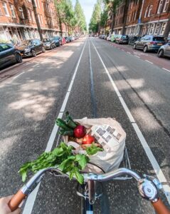 Abnehmen Psyche austricksen: Das Bild zeigt aus der Sichtweise einer Radfahrer*inn den Fahrradlenker mit einem Fahrradkorb voller gesunder Lebensmittel. Die Person fährt auf einer langen geraden Straße, ohne Autoverkehr.